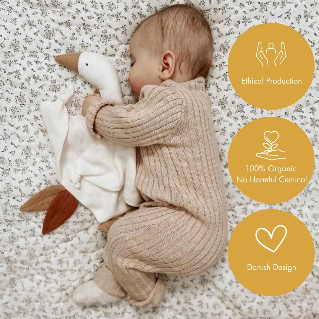 Dojenček v rjavem pletenem pajacu, ki spi na belem rjuhi in drži odejico za tolažbo v obliki goske.