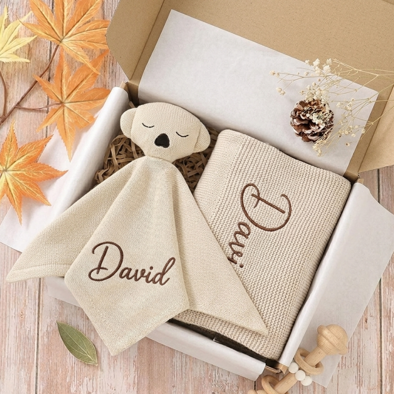 Personaliziran darilni set za dojenčka v bež barvi: bombažna odejica in ninica z vezenim imenom David, zapakirana v darilno škatlo.