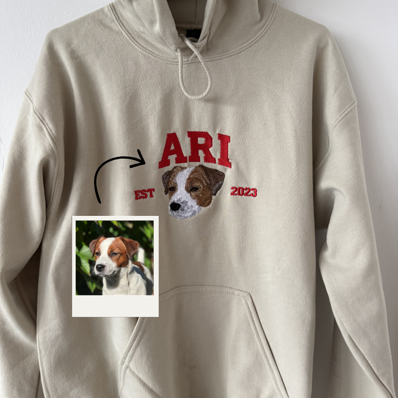 Personaliziran bež pulover s kapuco in vezenim portretom psa Jack Russell, napisom Ari ter letnico, izdelan po fotografiji stranke.
