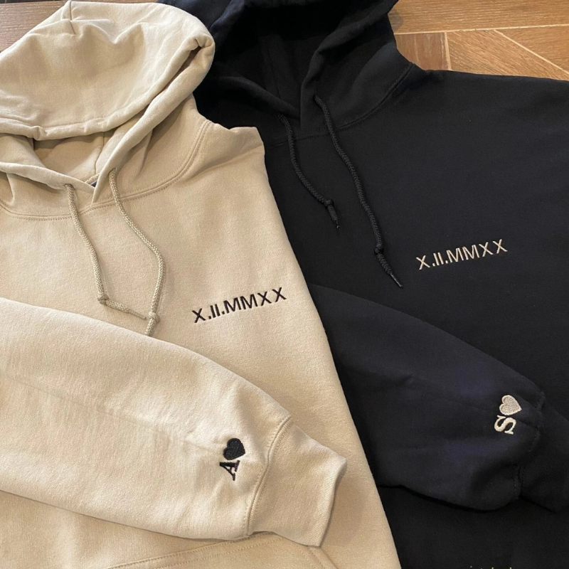 Ujemajoča se puloverja s kapuco v bež in črni barvi z vezenim datumom obletnice v rimskih številkah na prsih ter inicialkama A in S s srčkom na rokavu.